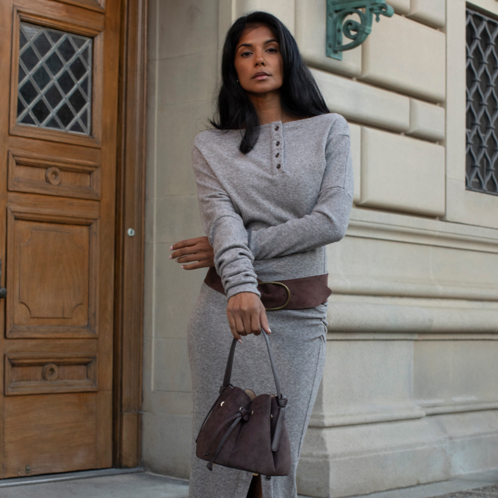 A woman stands outdoors in front of a stone building and wooden door, wearing a light gray long-sleeve henley top and matching gray skirt or dress, styled with a wide brown belt and carrying a brown handbag.