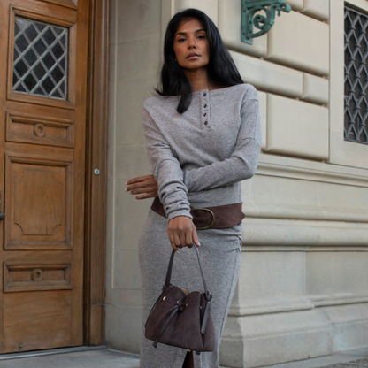 A woman stands outdoors in front of a stone building and wooden door, wearing a light gray long-sleeve henley top and matching gray skirt or dress, styled with a wide brown belt and carrying a brown handbag.