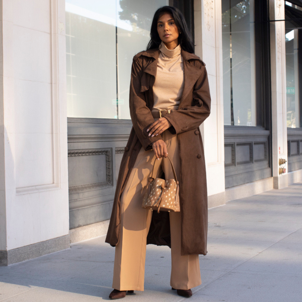 A woman stands on a city sidewalk wearing a long brown trench coat over a beige turtleneck and wide-leg pants, holding a fawn print bucket handbag.