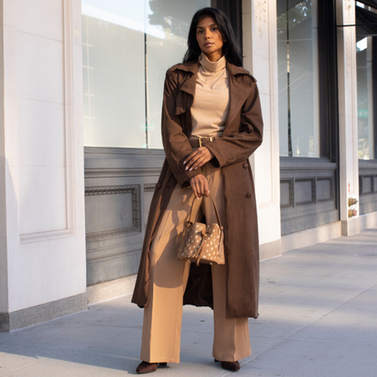 A woman stands on a city sidewalk wearing a long brown trench coat over a beige turtleneck and wide-leg pants, holding a fawn print bucket handbag.