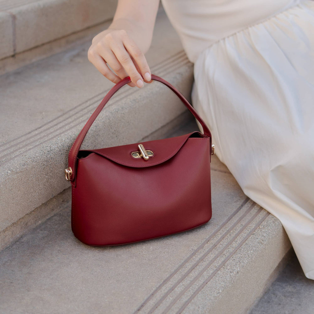 A burgundy leather handbag with a top handle and gold clasp, held by a person wearing a white dress while sitting on stone steps.