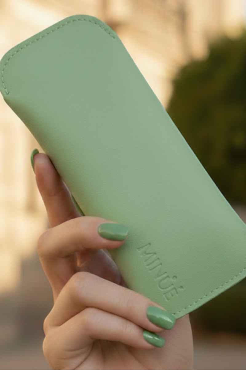 Hand holding a green glasses case with 'MINUE' branding outdoors.