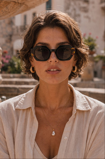 A woman with short brown hair wearing round black sunglasses, gold hoop earrings, and layered gold necklaces, dressed in a light beige button-up shirt. The background shows a softly blurred outdoor setting with stone steps and warm-toned buildings.