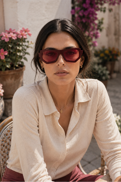 A woman wearing oversized dark red sunglasses and a cream ribbed knit polo shirt, seated in a wicker chair in an outdoor patio setting with potted flowers.