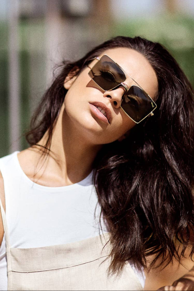 A woman with long dark hair wearing square sunglasses and a white sleeveless top leans against a glass wall, with her reflection visible on the left and a blurred outdoor background behind her.