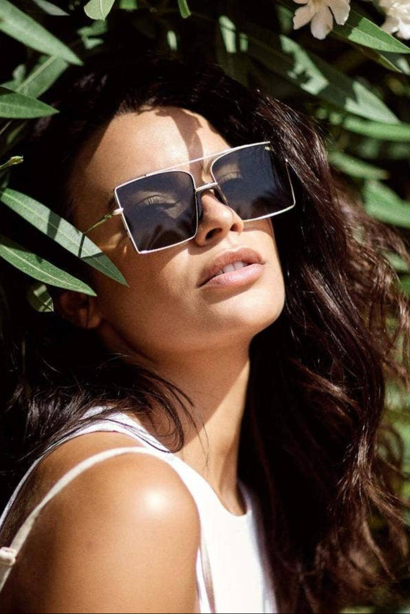 A woman wearing square sunglasses with a silver-toned metal frame and dark lenses, photographed outdoors against a background of green leaves and white flowers.