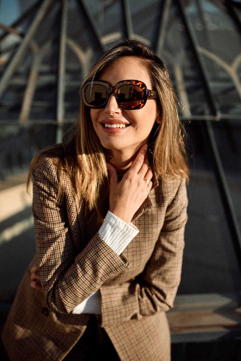 A woman wearing oversized tortoiseshell sunglasses and a brown plaid blazer, smiling in front of a modern glass architectural structure.
