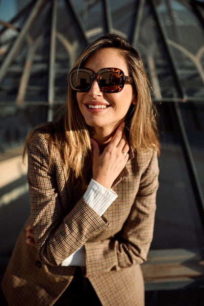A woman wearing oversized tortoiseshell sunglasses and a brown plaid blazer, smiling in front of a modern glass architectural structure.