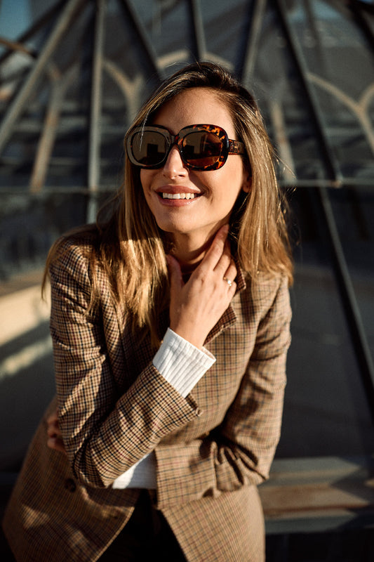 A woman wearing oversized tortoiseshell sunglasses and a brown plaid blazer, smiling in front of a modern glass architectural structure.