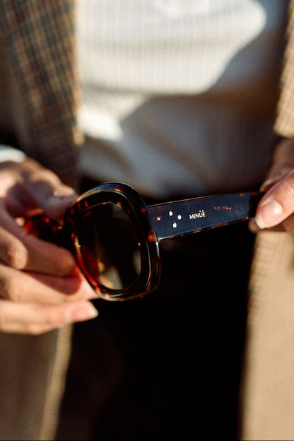 A close-up of a person holding a pair of dark sunglasses with tortoiseshell frames and a dark blue temple arm featuring white star details and the brand name 'smit'. The person is wearing a beige plaid blazer over a white top.