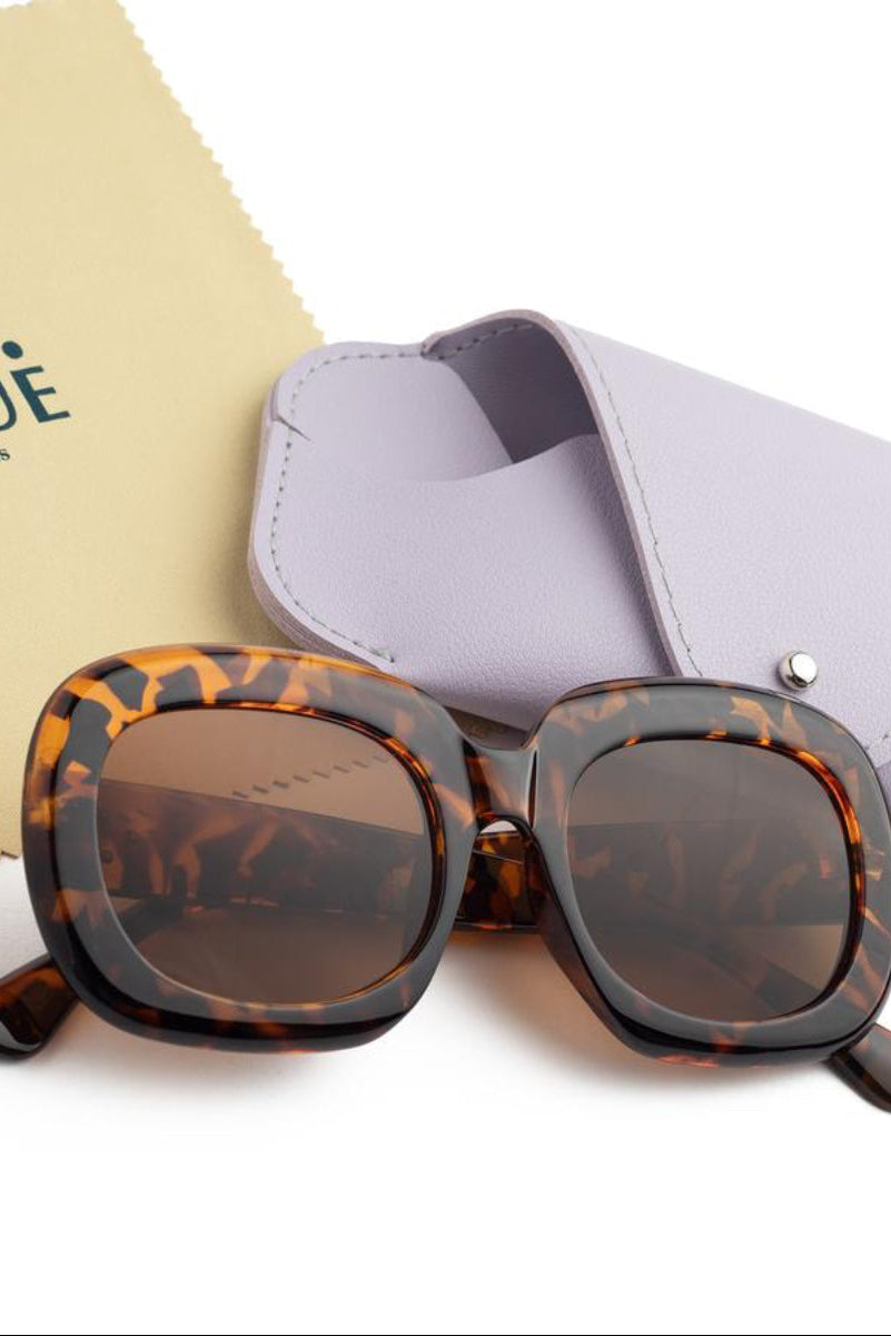 MINÜ Opticians square sunglasses with a brown tortoiseshell frame and dark lenses, displayed with a lavender case and a beige branded microfiber cloth on a white background.