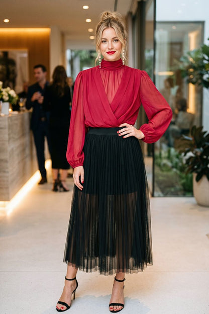 A woman models a two-piece outfit consisting of a deep red sheer long-sleeve mock-neck top and a black pleated midi skirt with a sheer tulle overlay, paired with black heeled sandals in a modern indoor lobby setting.