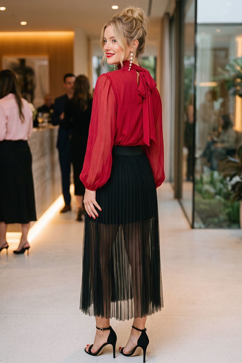 A woman stands in a modern indoor hallway wearing a bright red sheer long-sleeve blouse with a bow at the back of the neck, paired with a black pleated midi skirt and black high-heeled sandals.