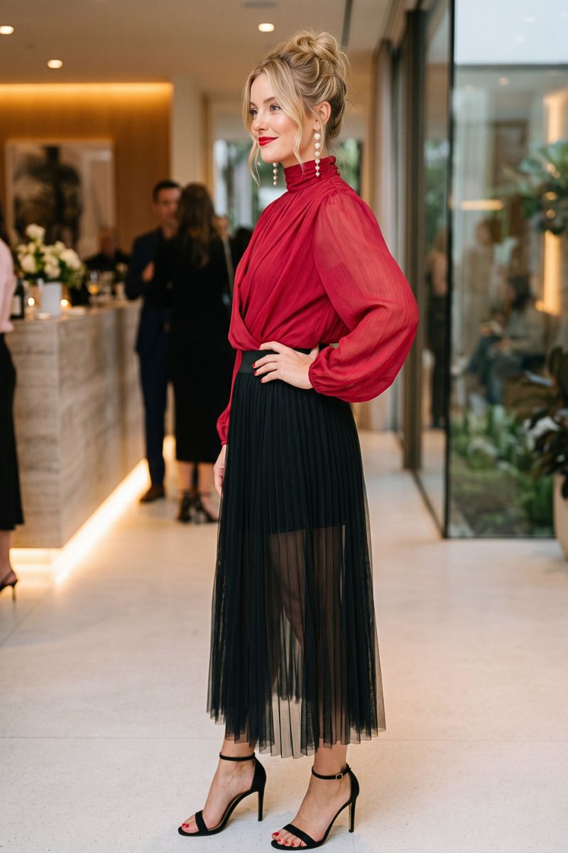 A woman stands in a modern indoor lobby wearing a sheer red high-neck blouse with long sleeves and a black pleated midi skirt with a sheer overlay, paired with black strappy heels.