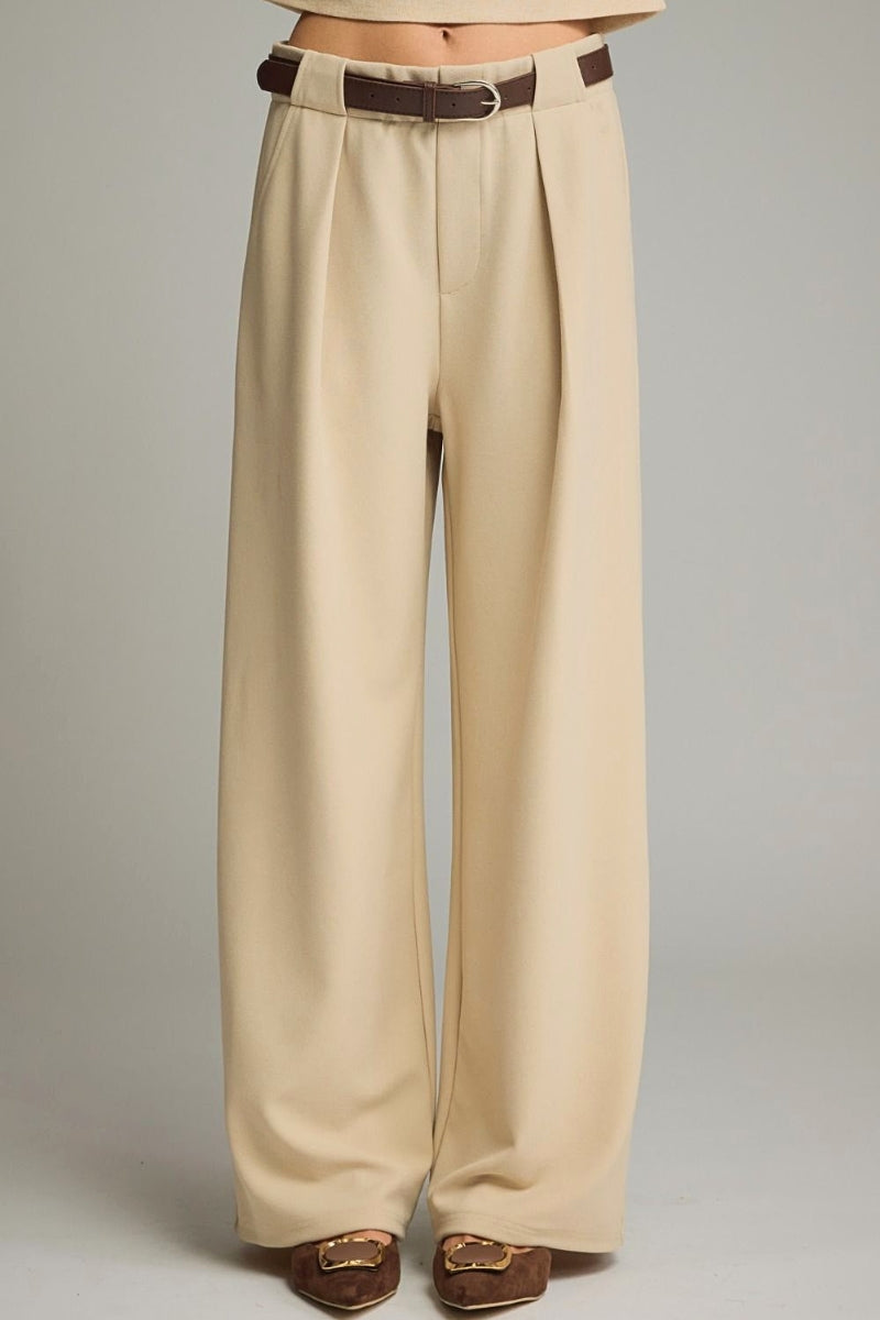 A person wearing high-waisted, wide-leg pleated trousers in a light beige/champagne color, styled with a dark brown belt and brown loafers against a neutral gray studio background.