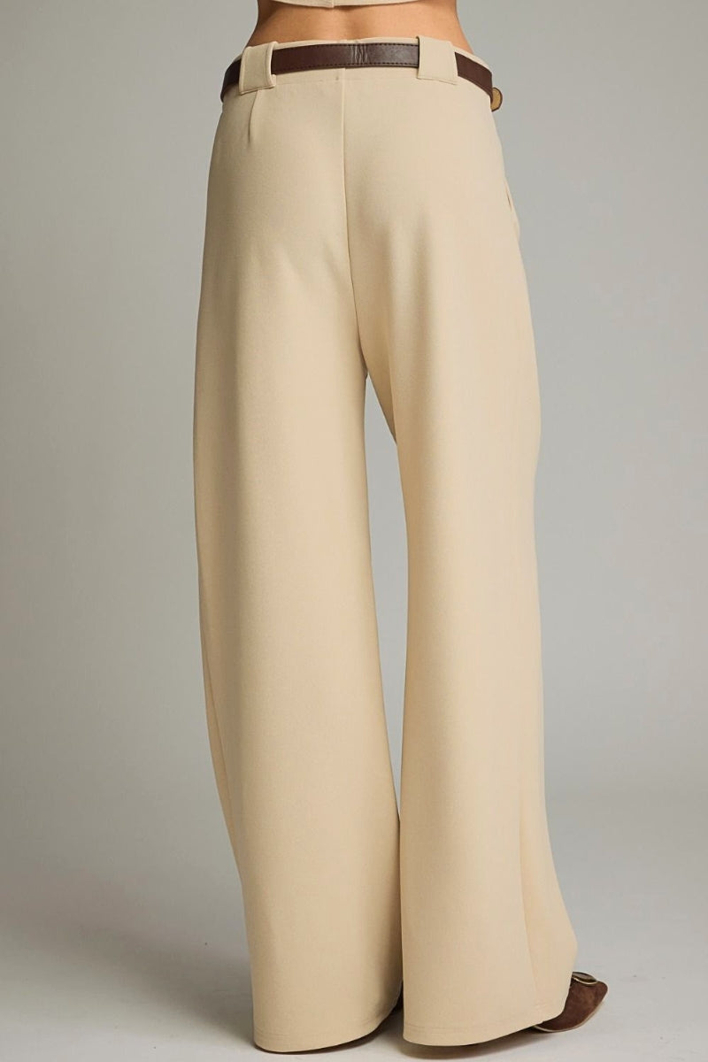 A person shown from the back wearing high-waisted, wide-leg trousers in a light beige color, styled with a dark brown belt and brown pointed-toe flats, against a neutral gray studio background.
