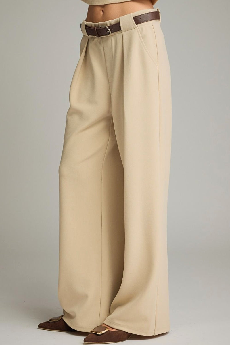 A person wearing high-waisted, wide-leg trousers in a light beige or cream tone, styled with a dark brown belt and brown pointed flats, against a neutral gray studio background.