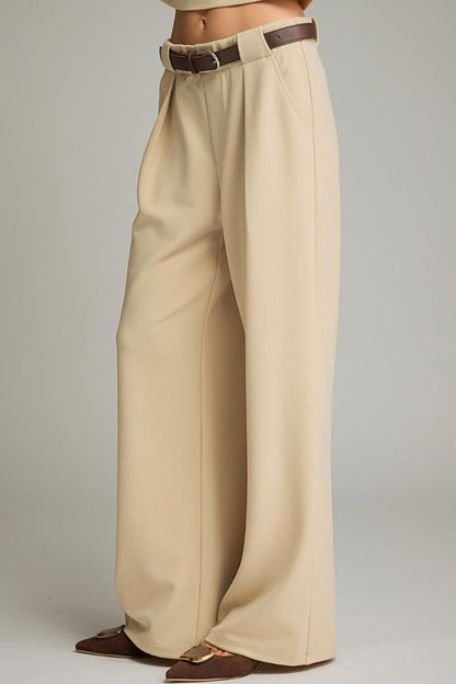 A person wearing high-waisted, wide-leg trousers in a light beige or cream tone, styled with a dark brown belt and brown pointed flats, against a neutral gray studio background.