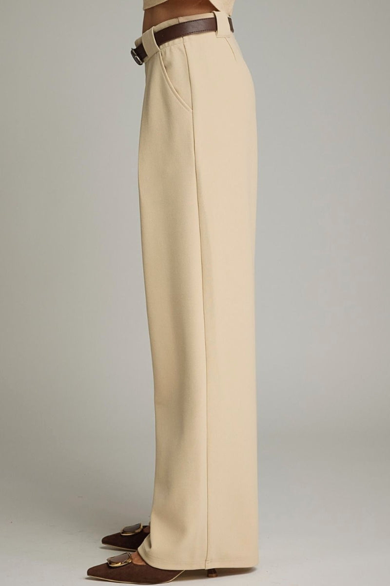 A side view of a model wearing high-waisted, wide-leg trousers in a light beige color, styled with a dark brown belt and brown pointed-toe flats against a neutral gray studio background.