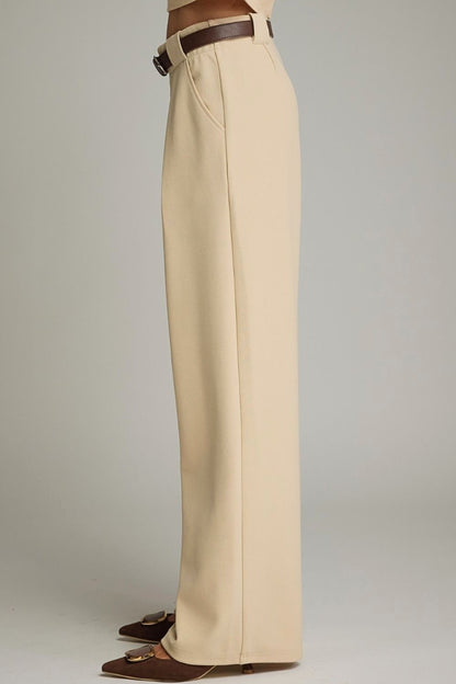 A side view of a model wearing high-waisted, wide-leg trousers in a light beige color, styled with a dark brown belt and brown pointed-toe flats against a neutral gray studio background.