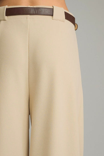 Close-up of a person wearing high-waisted cream-colored trousers with a dark brown leather belt, shown from the waist down against a plain gray background.