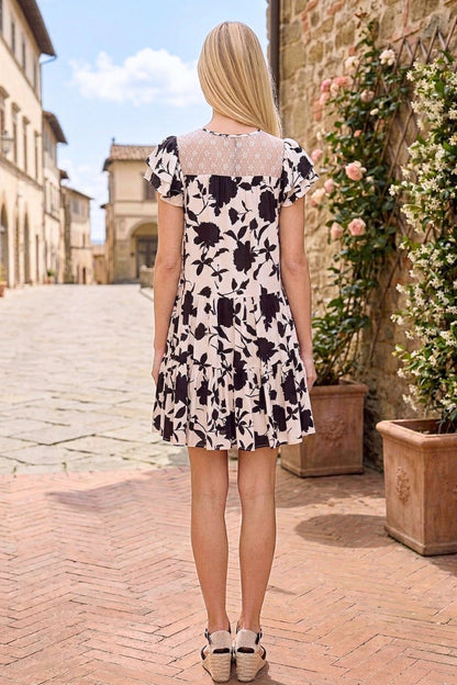 Woman in a floral dress standing on a stone path with buildings and plants in the background