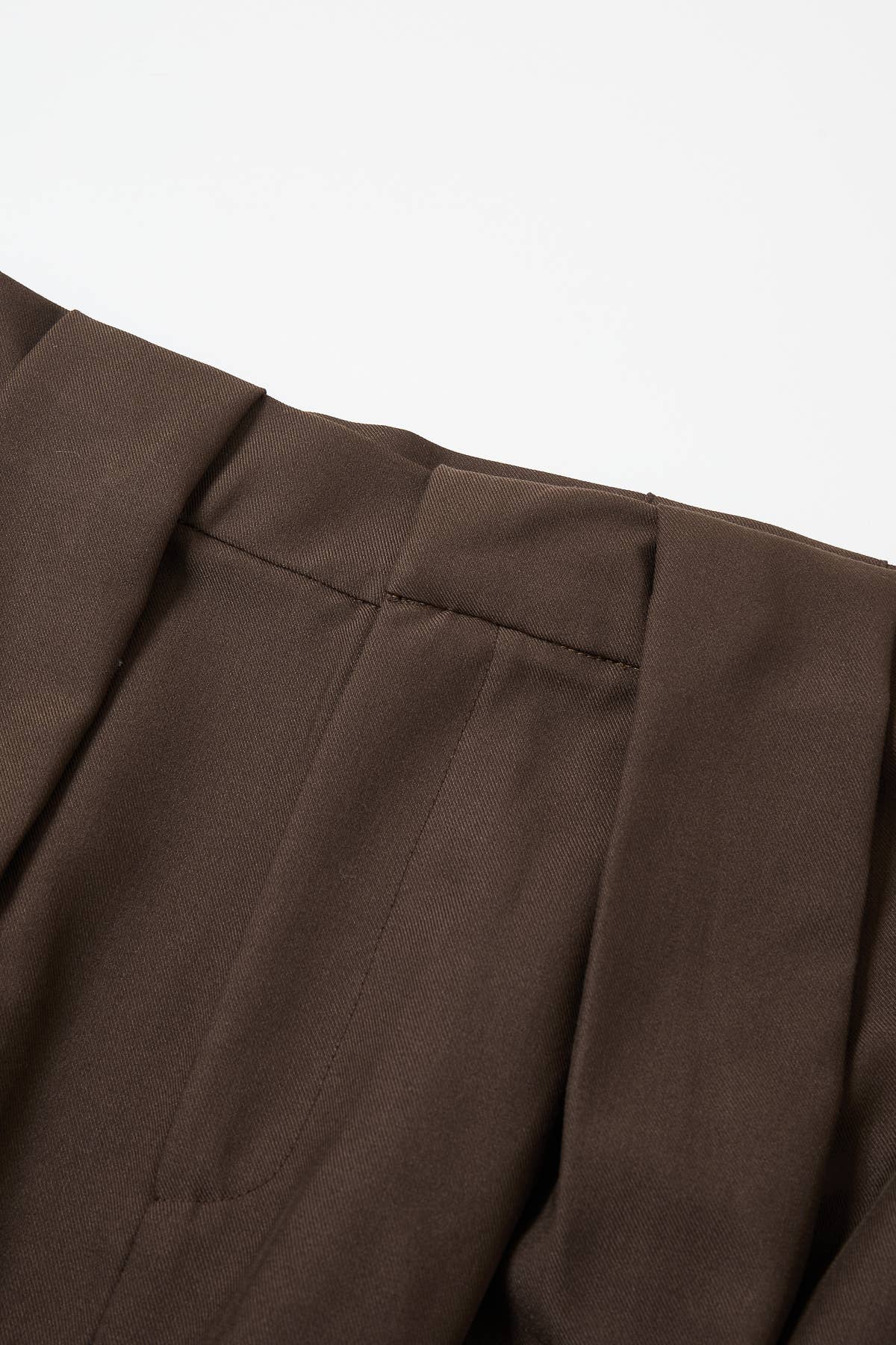 Close-up of dark brown pleated trousers showing the fabric texture and waistband stitching against a white background.