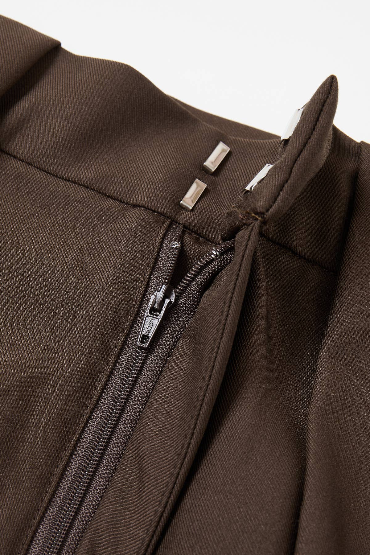 Close-up of a dark brown textured fabric garment with a black zipper and silver metal zipper pulls, set against a plain light background.