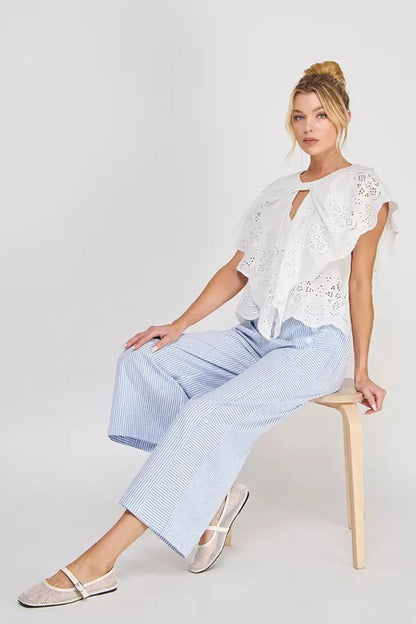 Woman sitting on a stool wearing a white lace top and light blue pants against a plain background