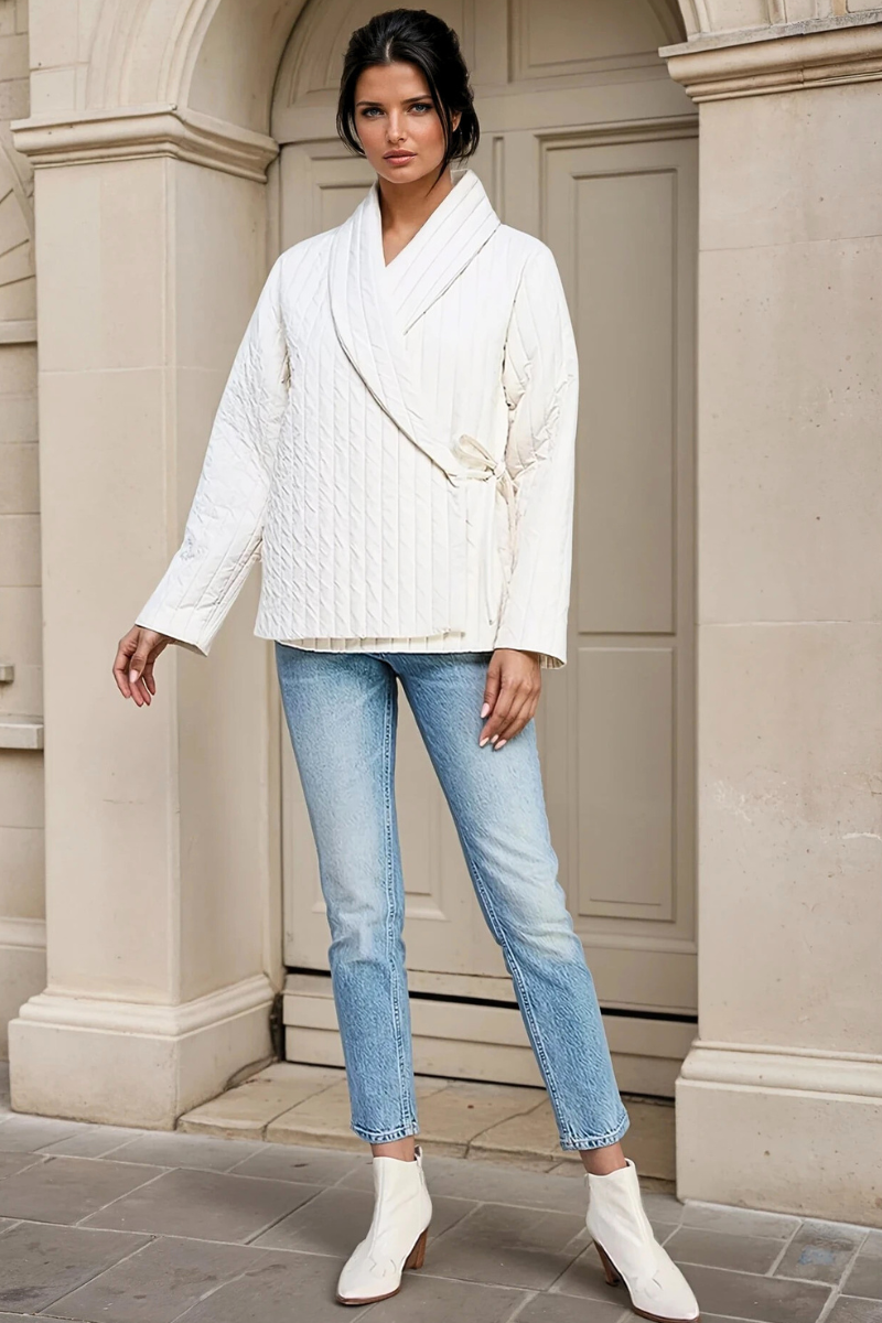 Woman wearing a white jacket, blue jeans, and white boots standing in front of a beige building.