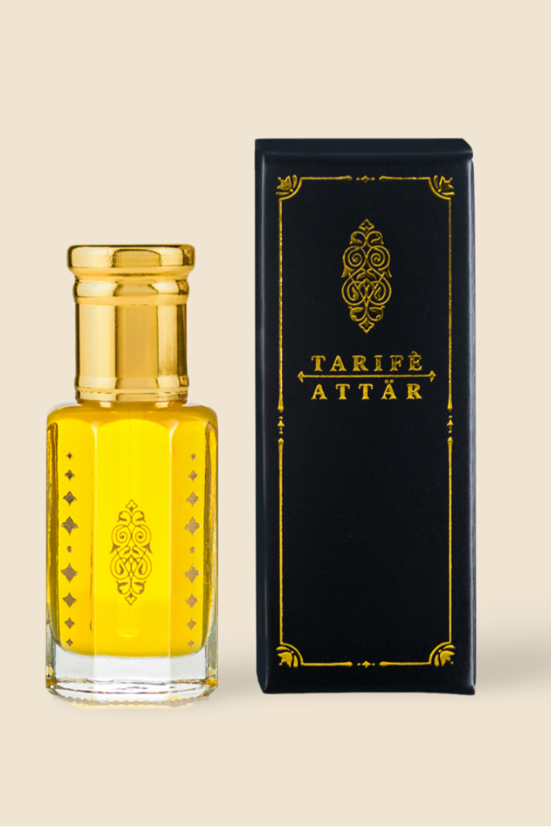 A bottle of Tarife Attâr perfume oil filled with golden-yellow liquid, standing next to its black and gold packaging box on a solid beige background.