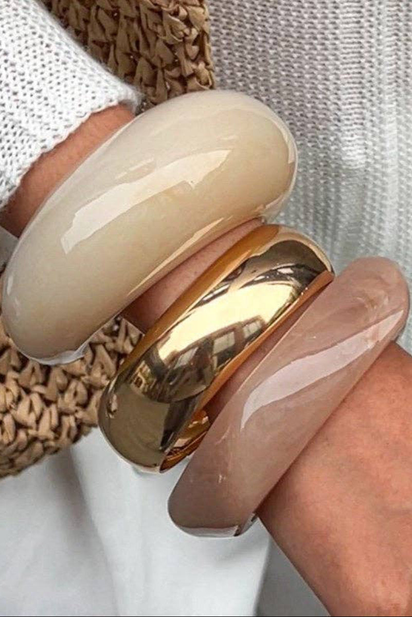 Close-up of a hand wearing two bangles, one beige and one gold, on a neutral background.