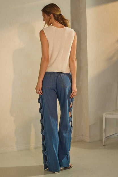 Woman wearing a sleeveless top and blue jeans standing against a plain wall.