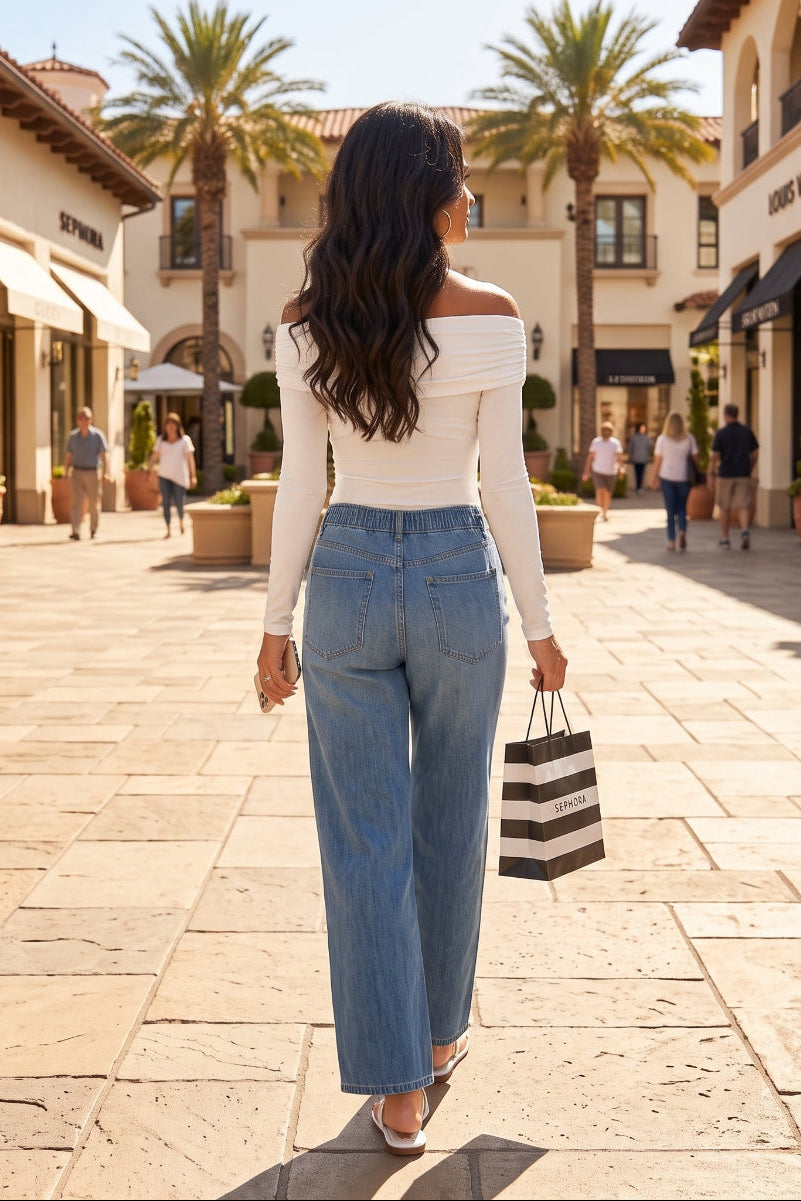 A woman seen from behind walking down a sunlit outdoor shopping plaza, wearing a white off-the-shoulder long-sleeve top and high-waisted wide-leg blue jeans, holding a black and white striped shopping bag.