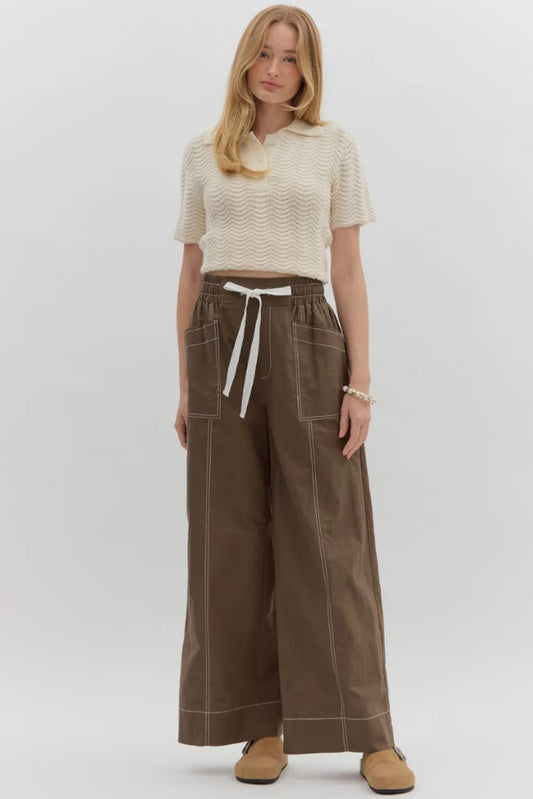 Woman wearing a cream top and brown wide-leg pants with a white belt on a white background