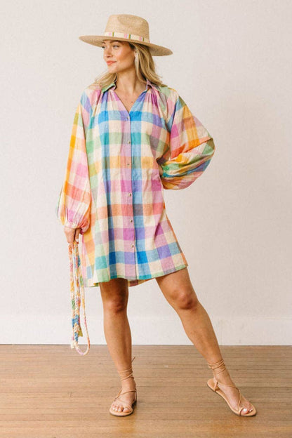Woman wearing a colorful plaid dress and straw hat on a wooden floor with a white wall background