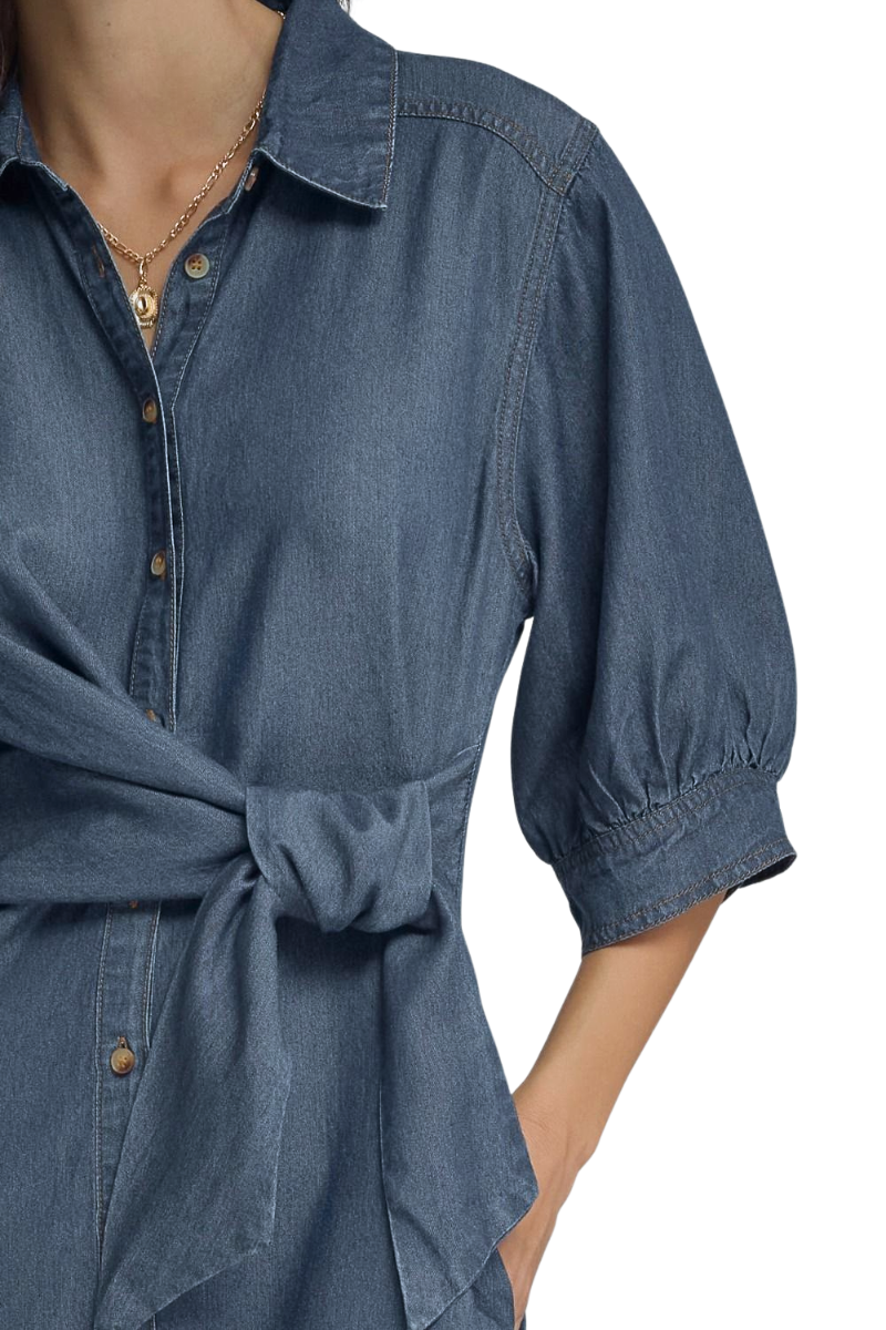A close-up of a person wearing a blue denim shirt dress with a tie waist and puff sleeves, shown against a plain white background.
