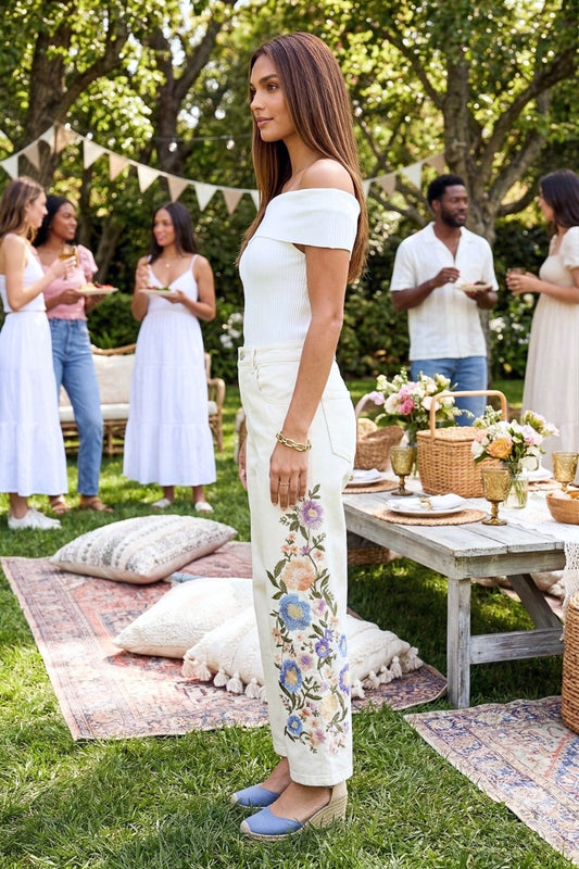 Woman in a white top and floral pants standing in a garden with people and a picnic setup.