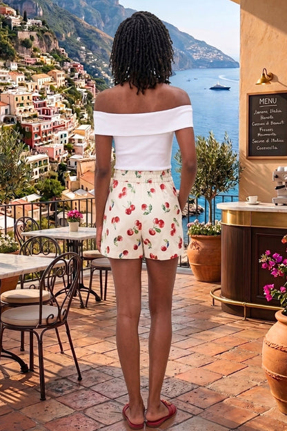 Woman in a white top and cherry print shorts standing on a scenic coastal street.
