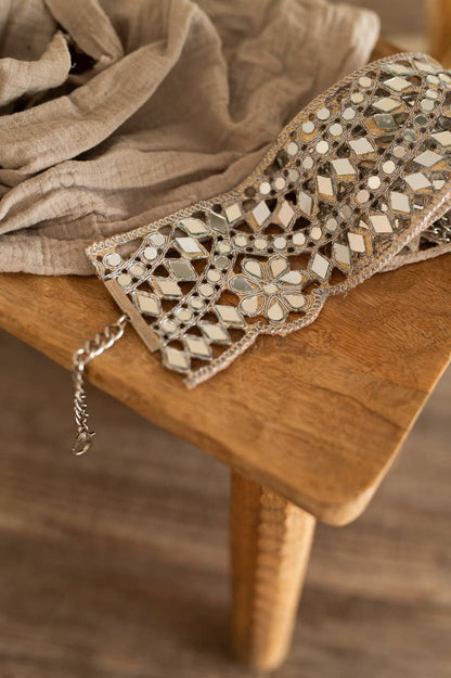 A close-up of a bohemian-style beaded belt with geometric mirror work and silver chain, resting on a rustic wooden table with a beige linen cloth.