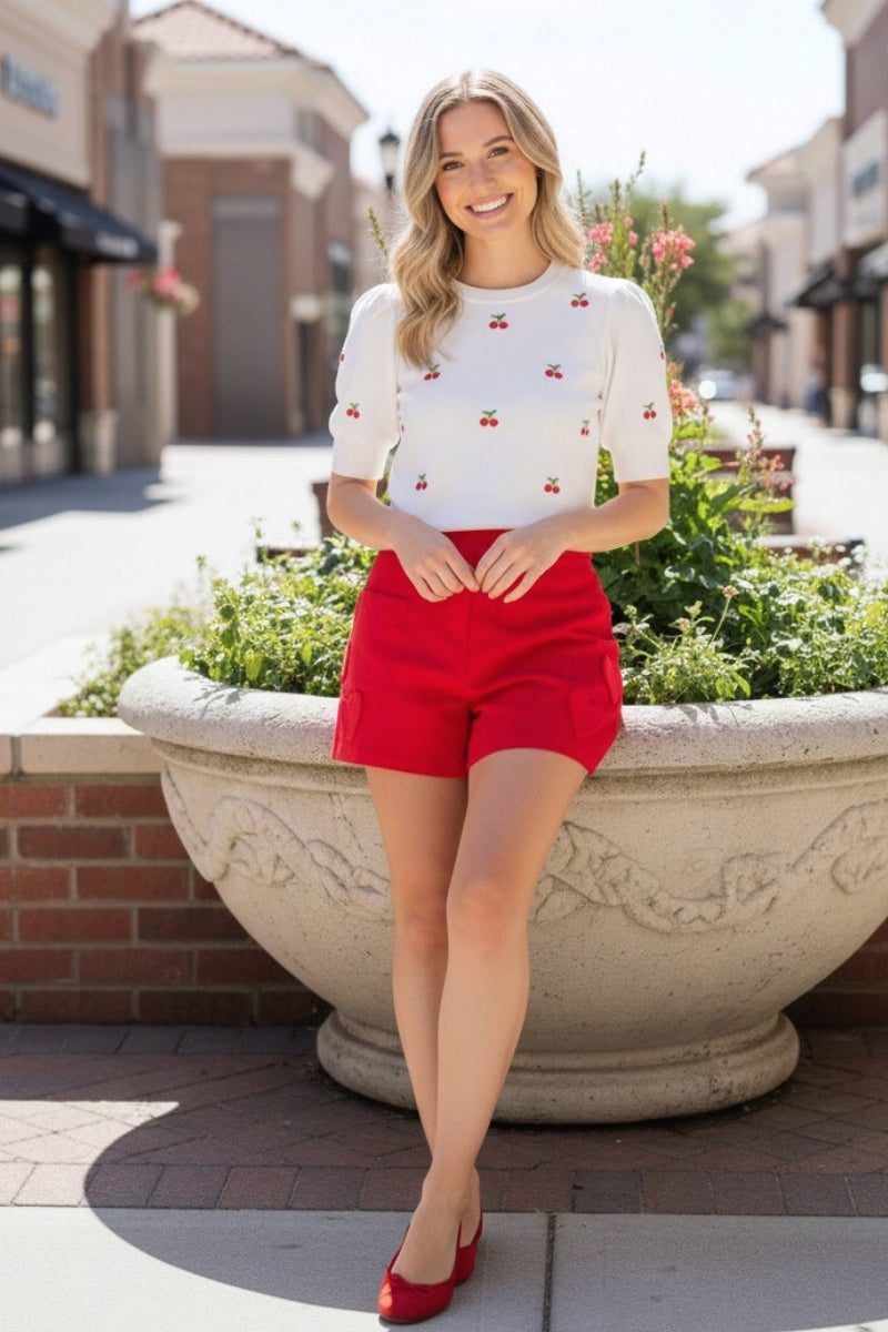 Woman in a white blouse with red accents and red shorts standing next to a large planter in an outdoor setting.