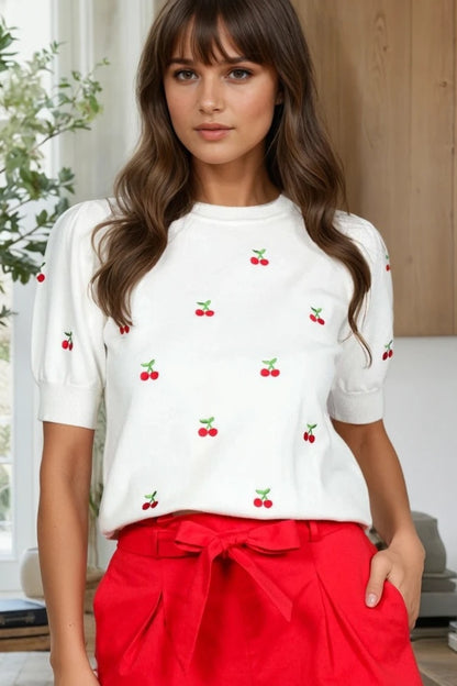 Woman wearing a white top with red cherry patterns and red pants indoors.