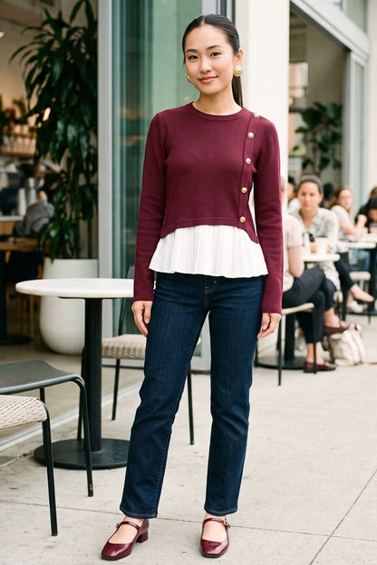 A woman stands outdoors wearing a burgundy long-sleeve cardigan with gold buttons and a white ruffled hem, paired with dark blue jeans and burgundy flats.