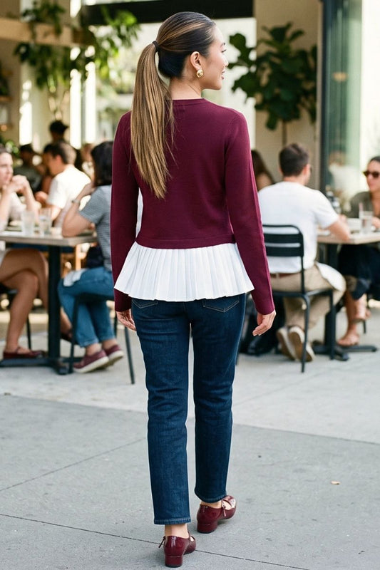 A woman viewed from behind wearing a burgundy long-sleeve top with a white pleated peplum hem, dark blue jeans, and burgundy flats, walking in an outdoor cafe setting.