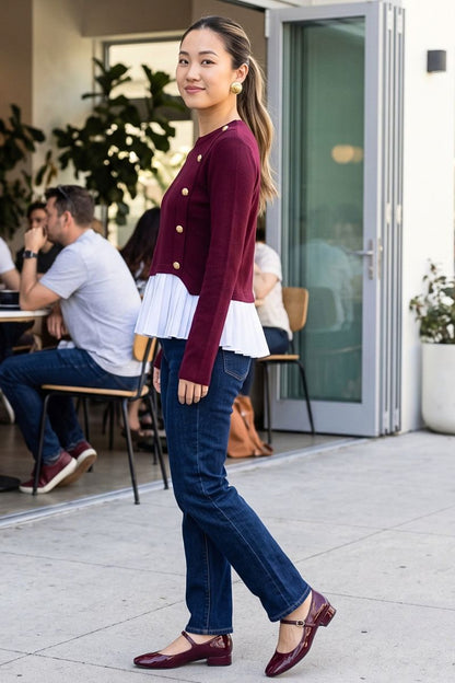A woman walking outdoors in front of a cafe, wearing a burgundy velvet cardigan with gold buttons and a white ruffled hem, paired with dark blue jeans and burgundy Mary Jane shoes.