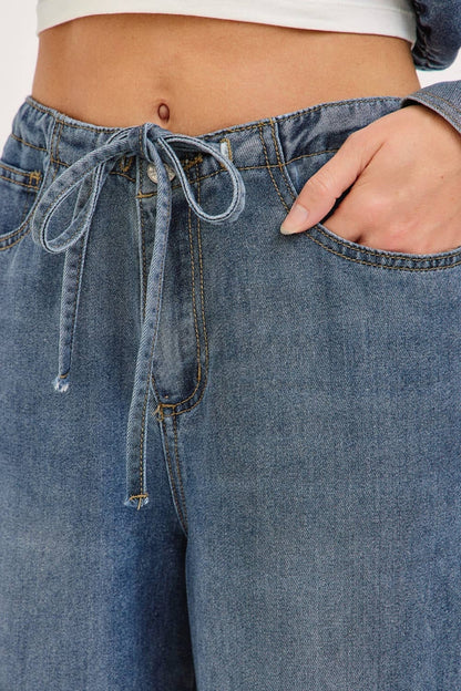 Close-up of a person wearing blue denim jeans with a drawstring waistband, showing the front pocket, side pocket, and hand in the pocket.