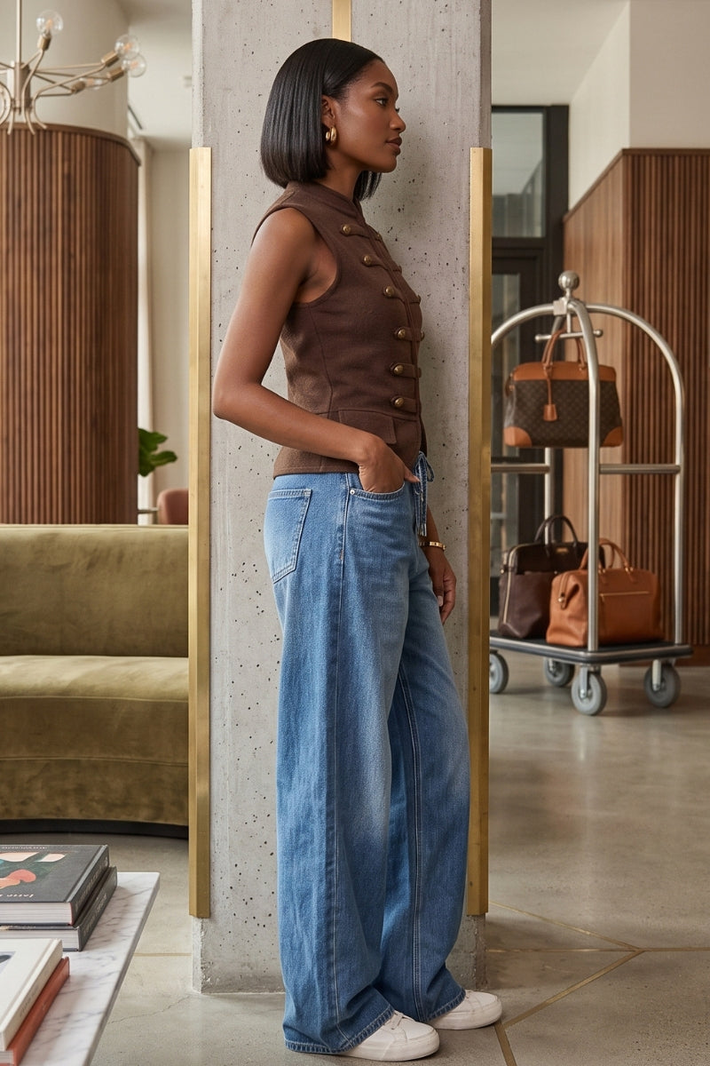 A woman stands in profile wearing a sleeveless brown vest with decorative buttons, wide-leg blue jeans, and white sneakers in a modern interior setting with a concrete pillar and luggage cart in the background.