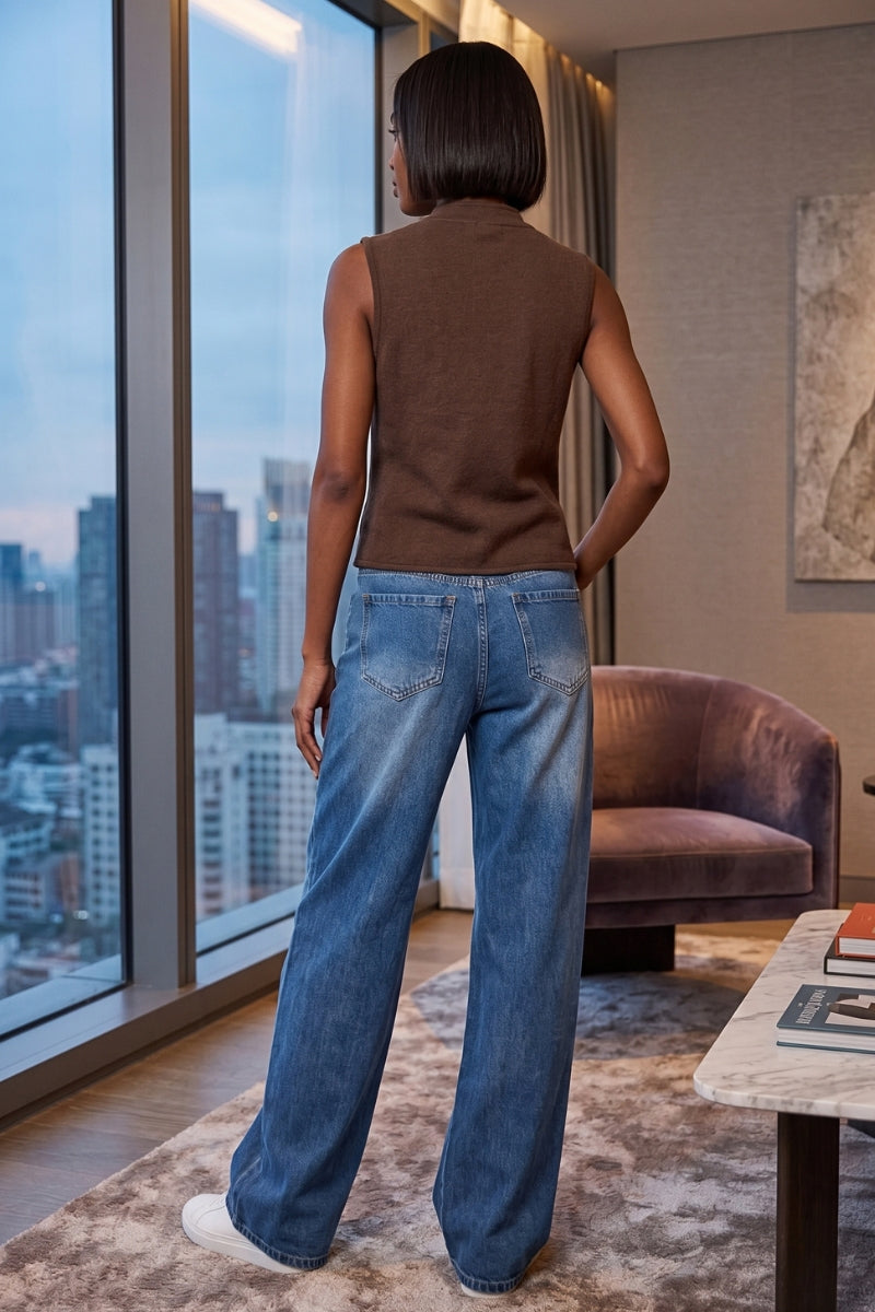 A woman stands with her back to the camera in a modern room, wearing a brown sleeveless knit top and wide-leg blue jeans, looking out a large window with a city skyline view.