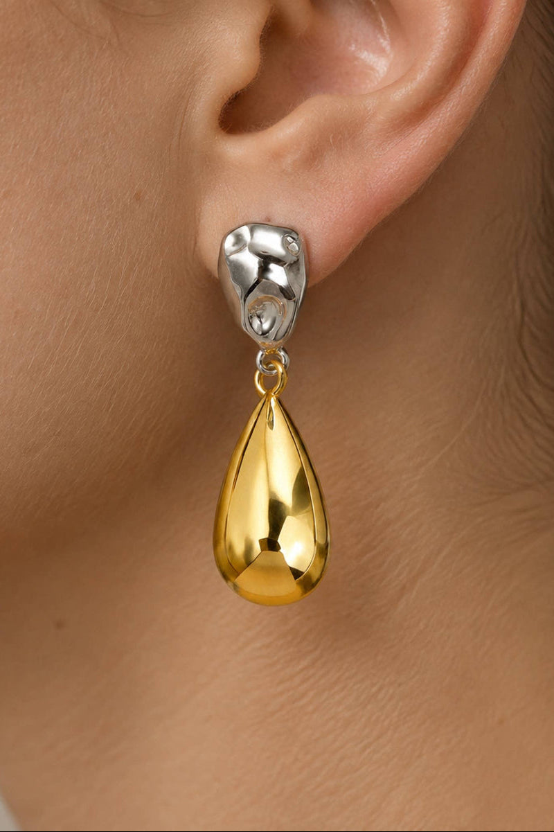 A close-up of a person's ear wearing a teardrop-shaped drop earring with a polished silver top and a shiny gold lower drop.
