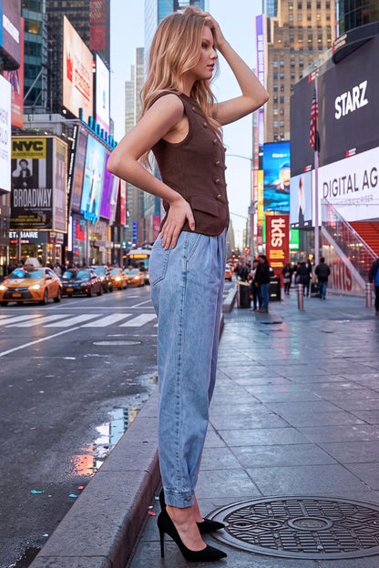 A woman stands on a city sidewalk in Times Square, wearing a brown sleeveless button-front top, light blue wide-leg jeans, and black high heels. Large billboards and street activity are visible in the background.